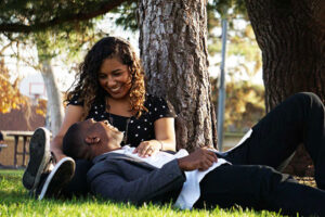 Male and female couple sitting under a tree, mans head is in womans lap and they're laughing together.